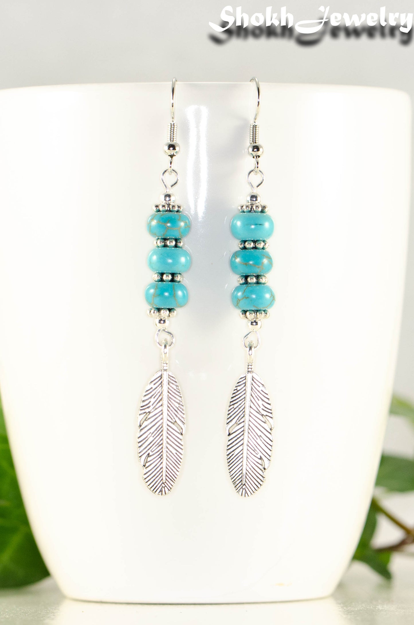 Statement Tibetan Silver Feather And Turquoise Howlite Earrings displayed on a coffee mug.