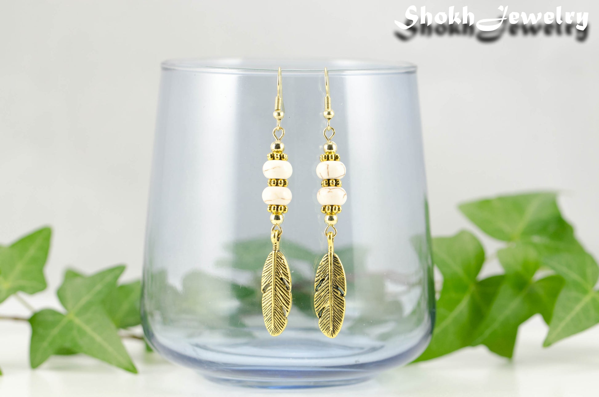 Statement White Howlite And Gold Feather Earrings displayed on a glass.