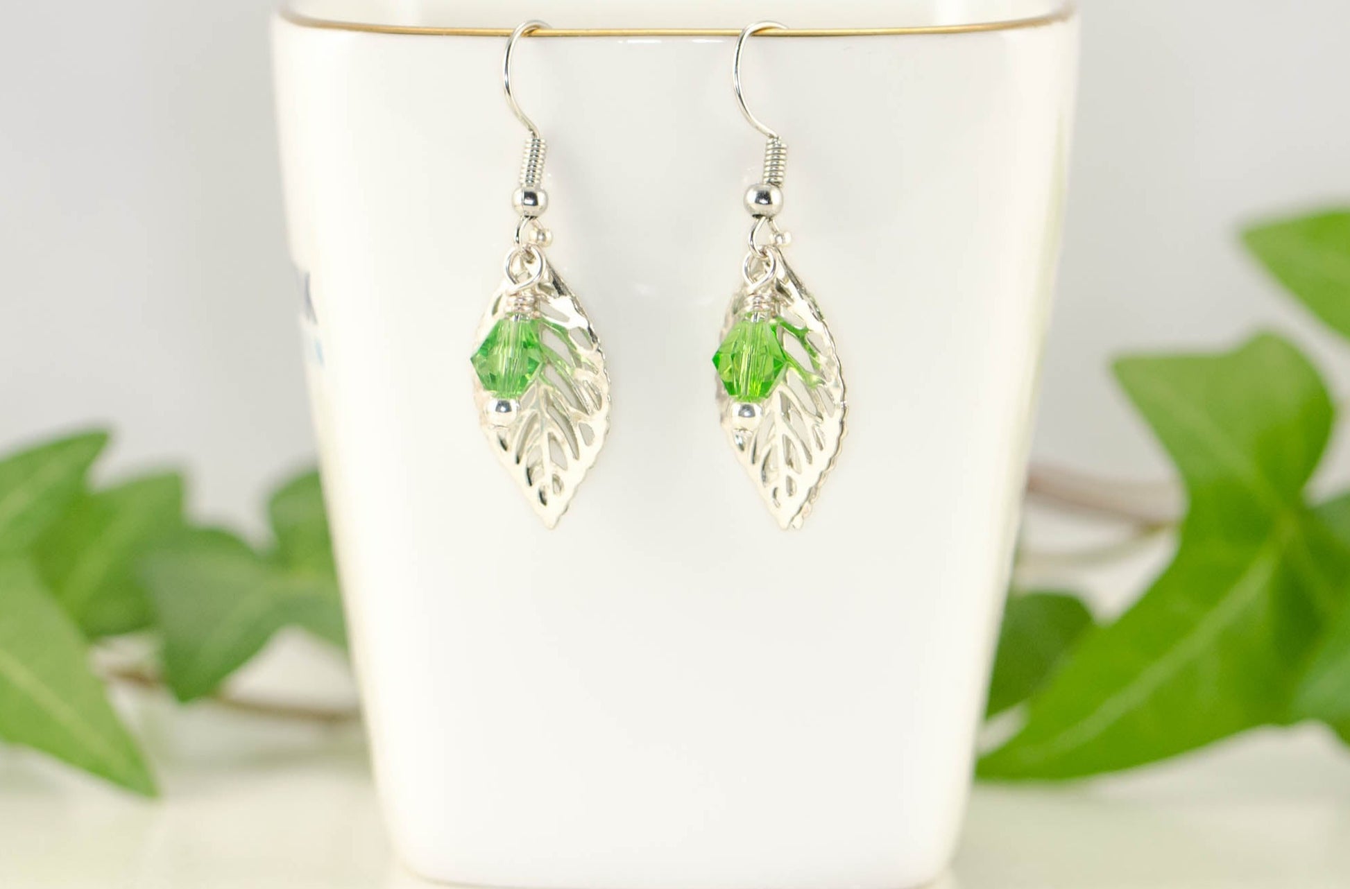 Filigree Silver Leaf Charm and Green Glass Crystal Earrings displayed on a tea cup.