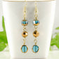 Gold and Patina Blue Glass Bead Dangle Earrings displayed on a tea cup.