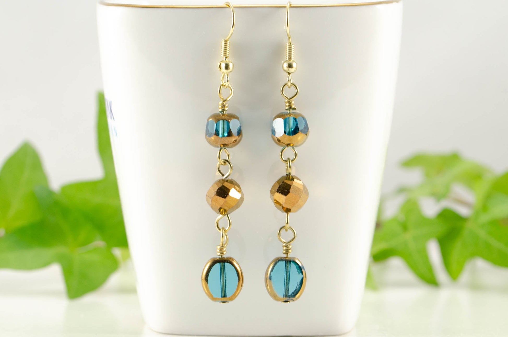 Gold and Patina Blue Glass Bead Dangle Earrings displayed on a tea cup.
