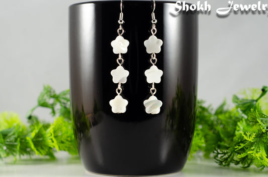 Long White Seashell Flower Earrings displayed on a black mug.
