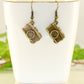 Antique Bronze Camera Charm Earrings displayed on a tea cup.