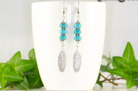 Statement Tibetan Silver Feather And Turquoise Howlite Earrings displayed on a coffee mug.