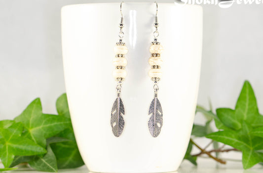 Statement White Howlite And Tibetan Silver Feather Earrings displayed on a coffee mug